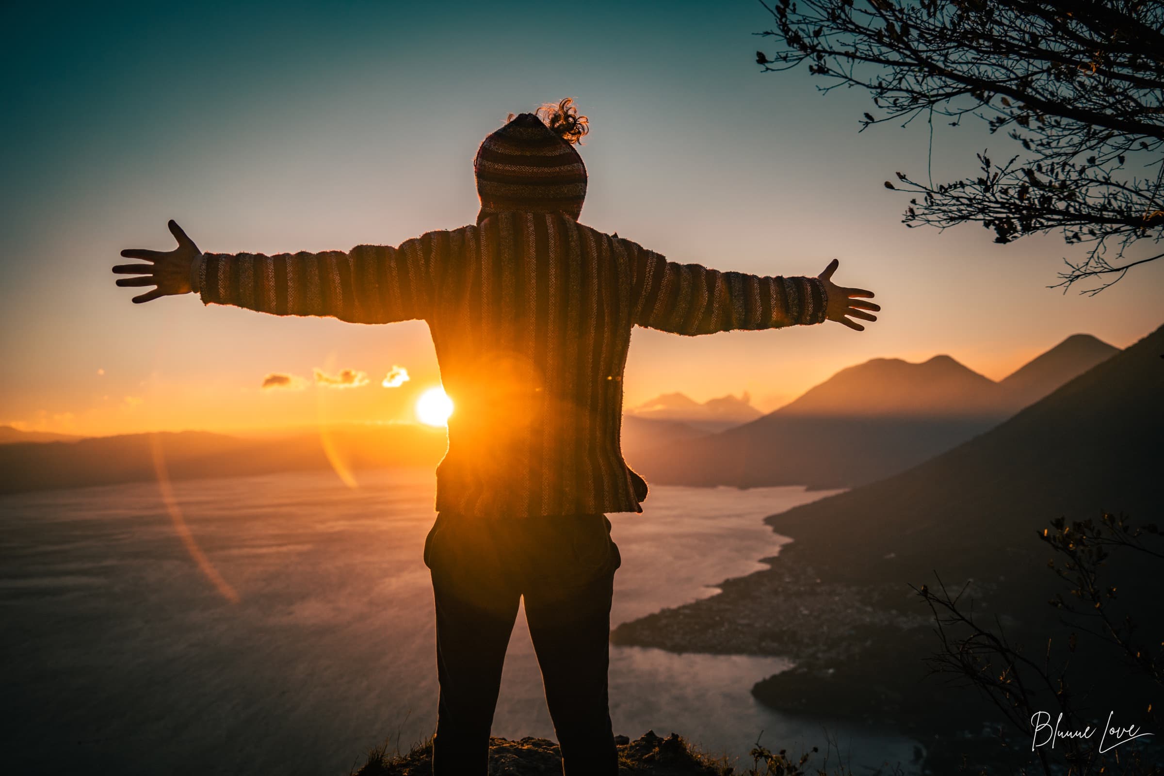 Joshua Sting standing with arms spread wide, facing a sunset over mountains and a lake